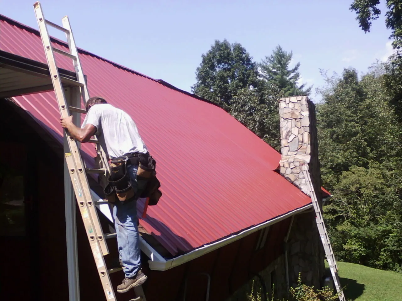 Roofer on a ladder beside a red metal roof and stone chimney — Soffit & Fascia Repair by Roofing Brothers in Austell